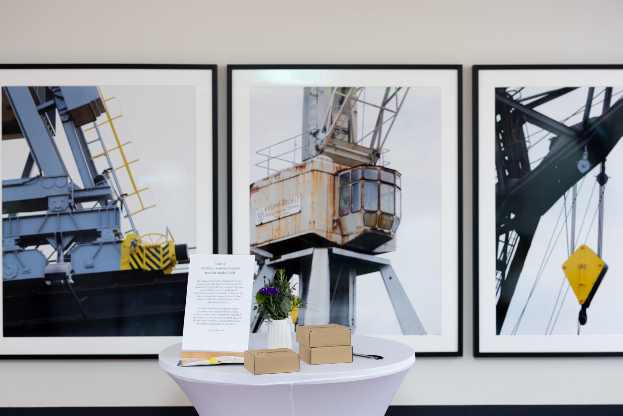 White standing table with product boxes and flowers in front of three framed harbour crane photographs.