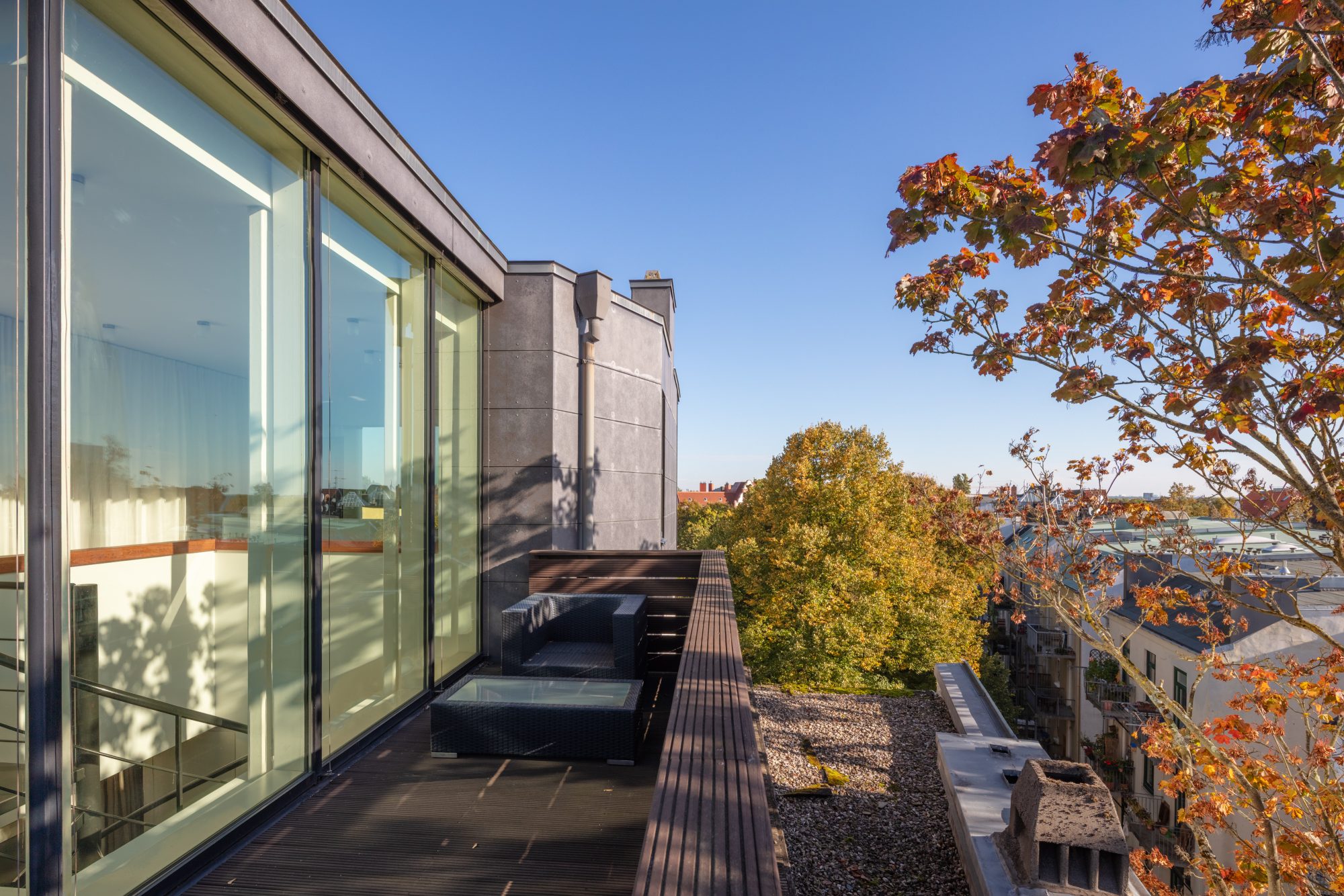 Schmale Dachterrasse mit Sitzmöbeln entlang der Glasfassade, umgeben von herbstlichen Bäumen und Hamburger Dächern.