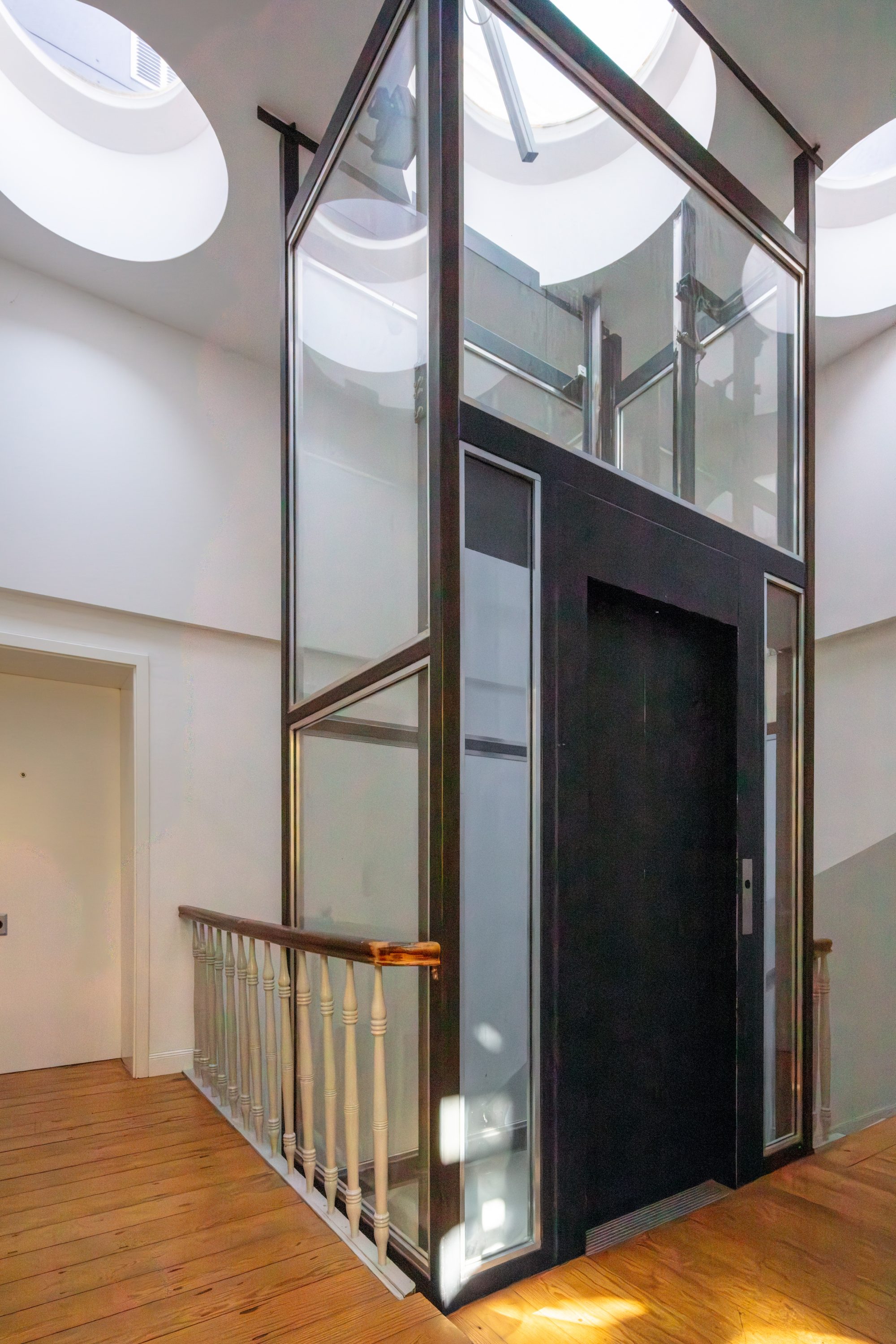 Verglaster Aufzugsschacht im Treppenhaus unter runden Oberlichtern, neben einem weißen Geländer und Holzboden.