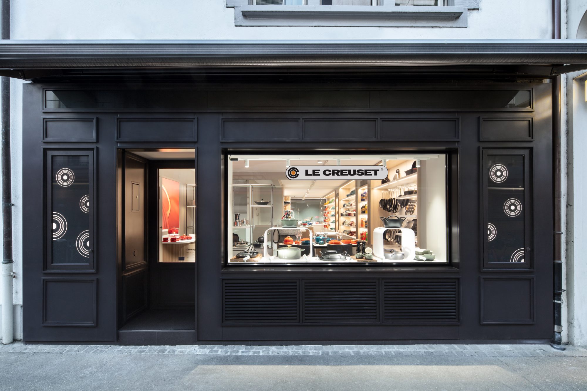 Schaufenster und Eingang der Le Creuset Signature Boutique in Basel mit beleuchteter Kochgeschirrpräsentation hinter einer dunklen Fassade.