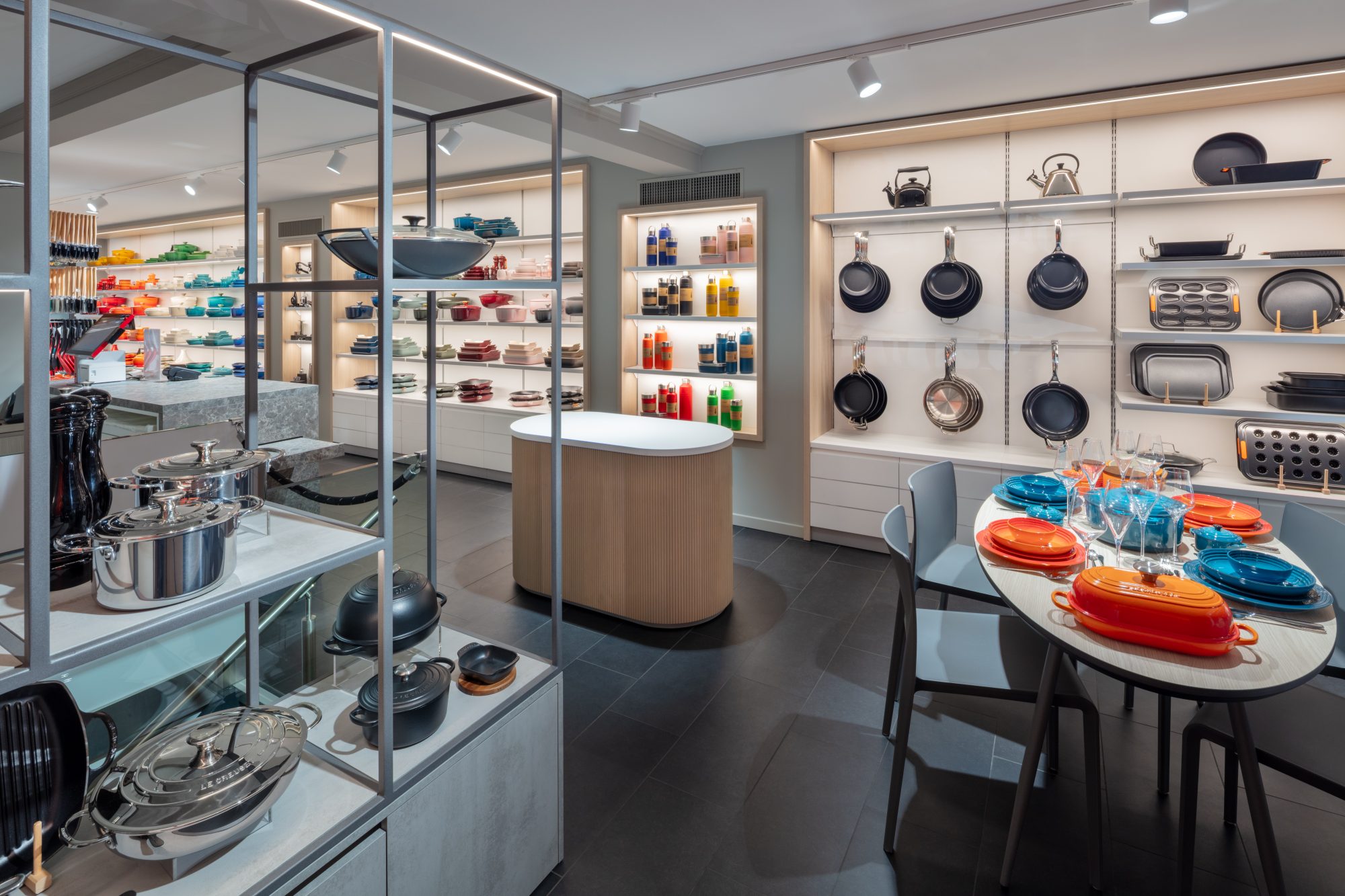 Präsentationsbereich der Le Creuset Boutique Basel mit Pfannen und Backformen an der Wand, Edelstahlkochgeschirr und einem gedeckten Tisch.
