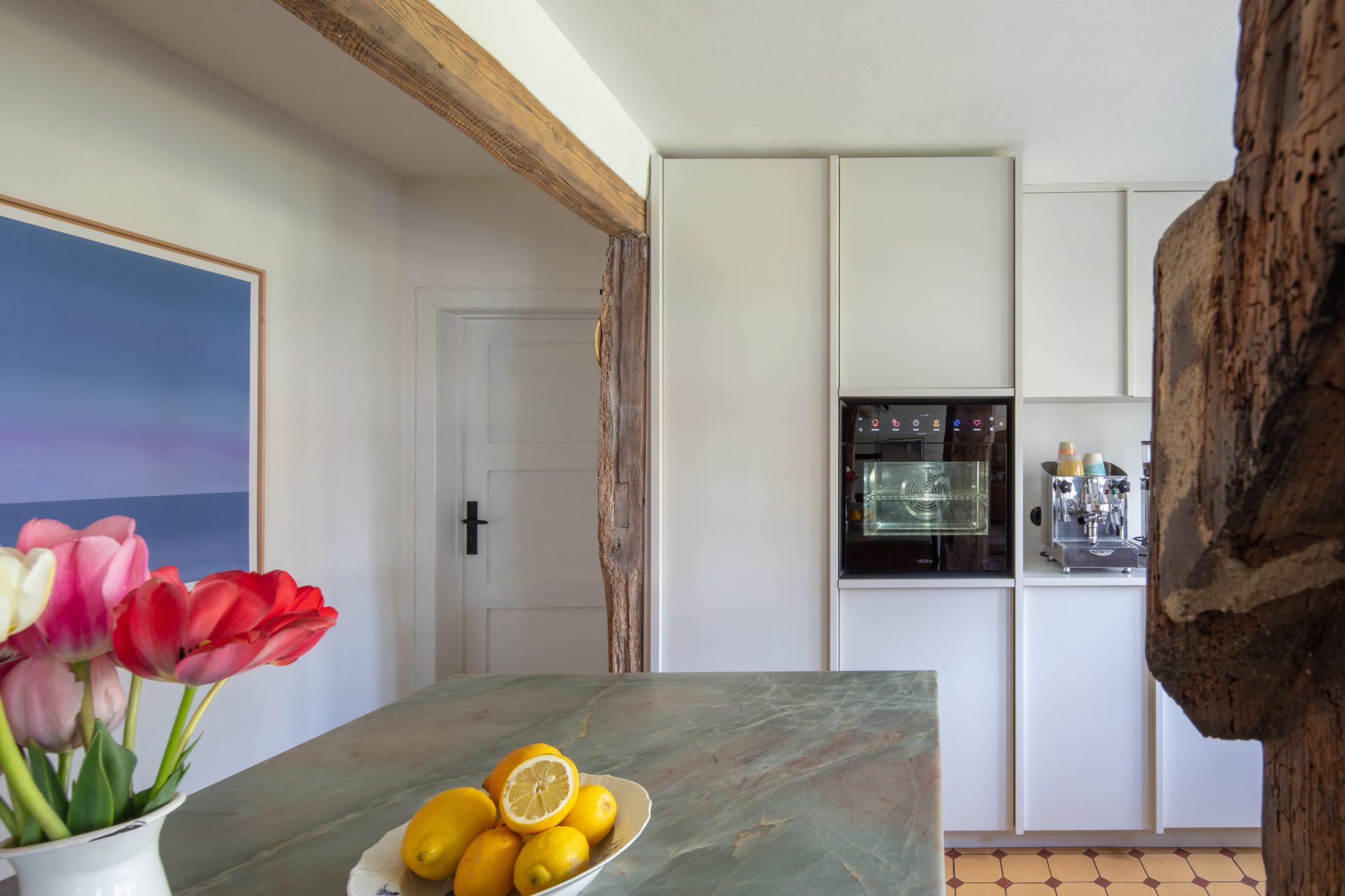 Helle Einbauschränke mit Backofen und Kaffeemaschine hinter der Kücheninsel, mit Tulpen und Zitronen im Vordergrund.