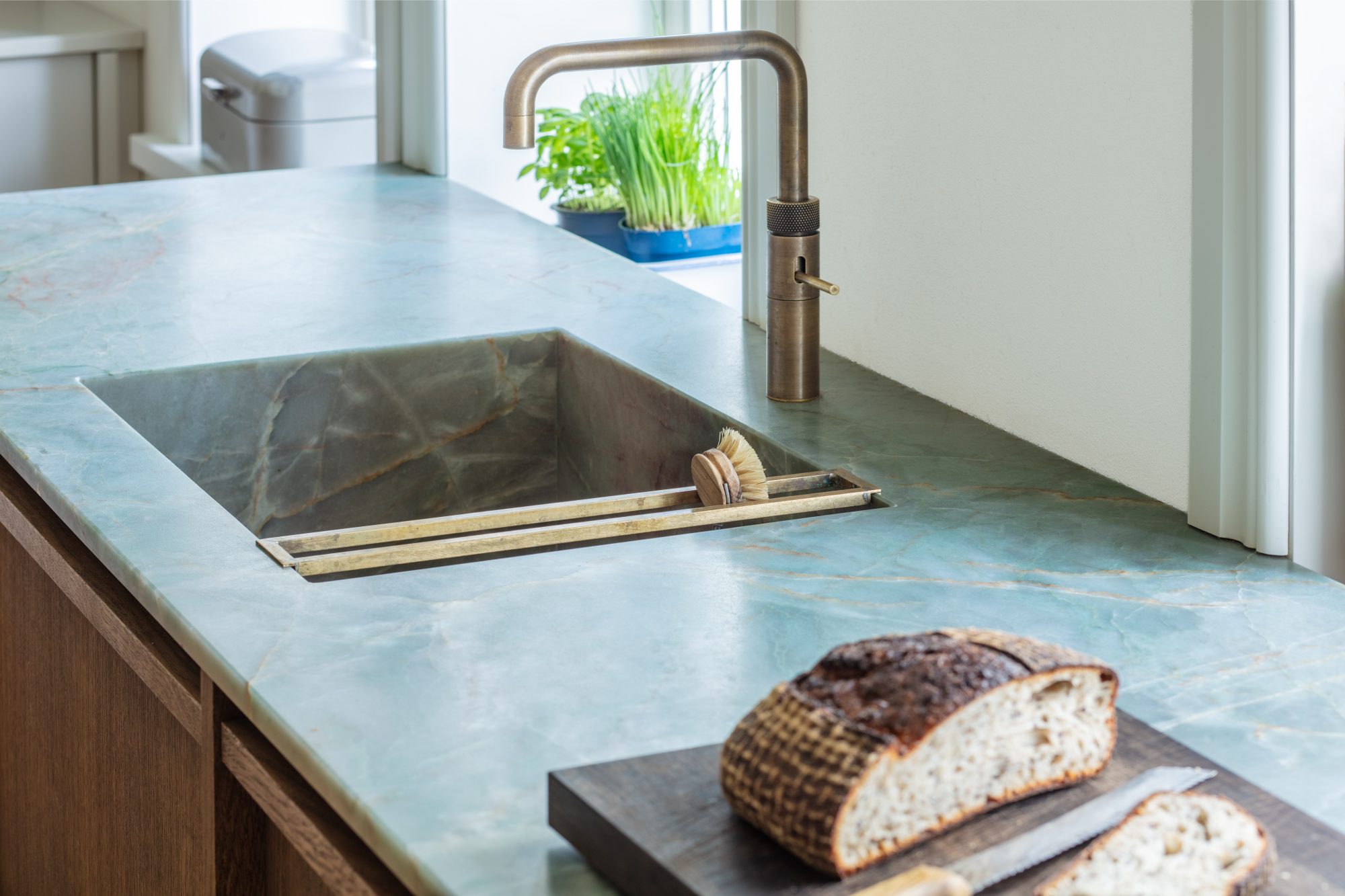 Detail der rechteckigen Spüle in der graugrünen Arbeitsfläche mit gebogener Armatur und Brot auf einem Schneidebrett.